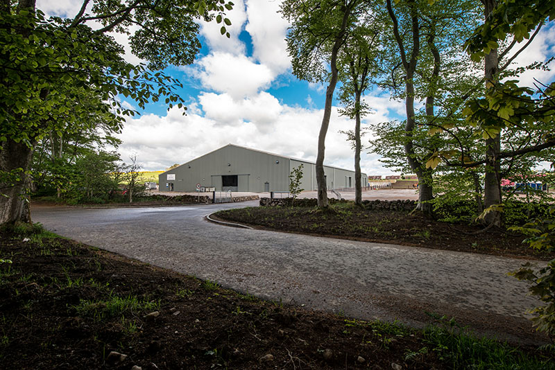 Curved road leading to an industrial warehouse, framed by green trees under a cloudy sky