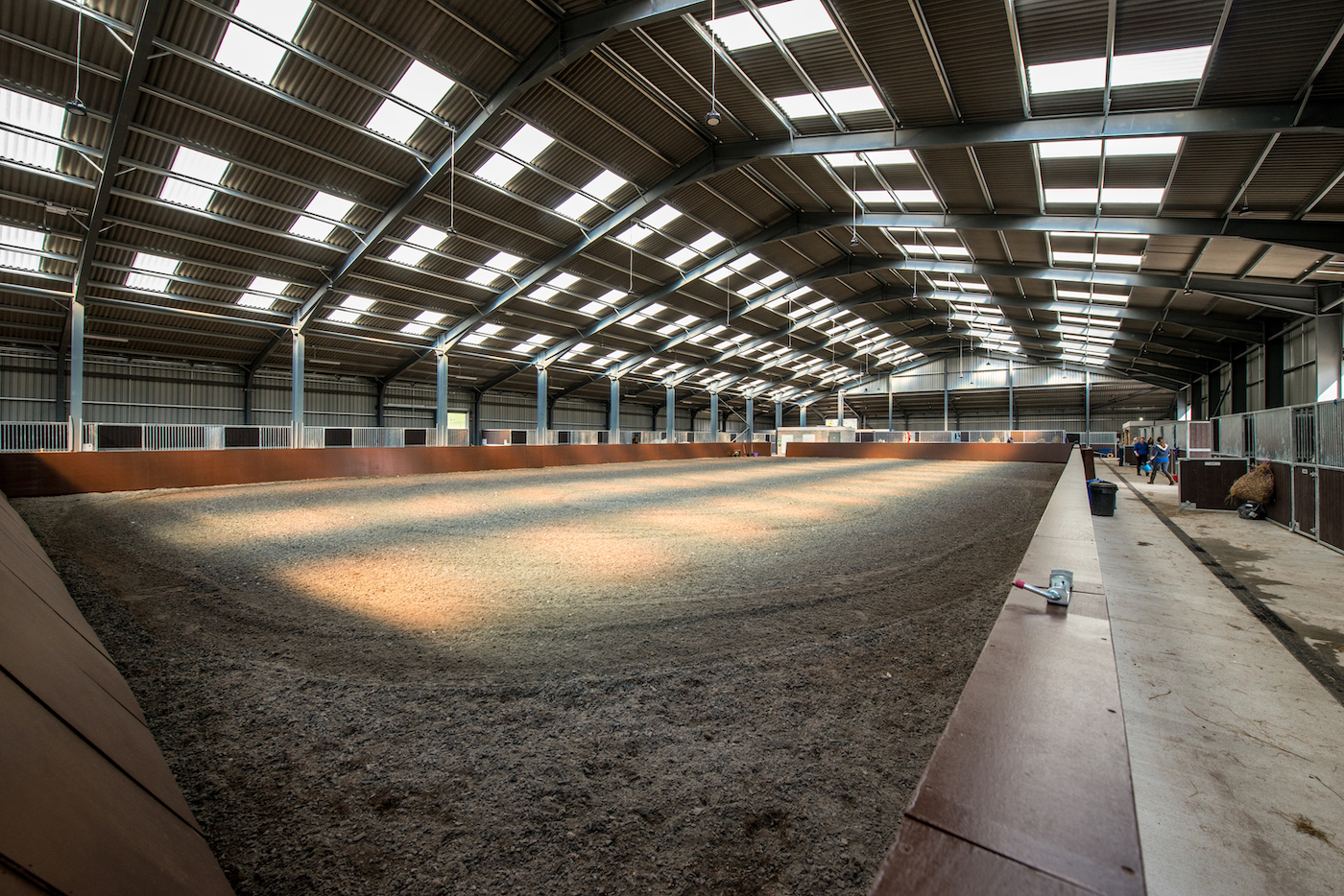 Indoor horse riding arena with a sandy floor and a high ceiling, sunlight streaming through skylights